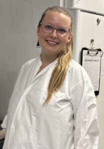 Smiling lab technician wearing safety glasses, gloves, and a white lab coat stands beside a Beckman Coulter centrifuge in a laboratory, with large freezers and equipment in the background.